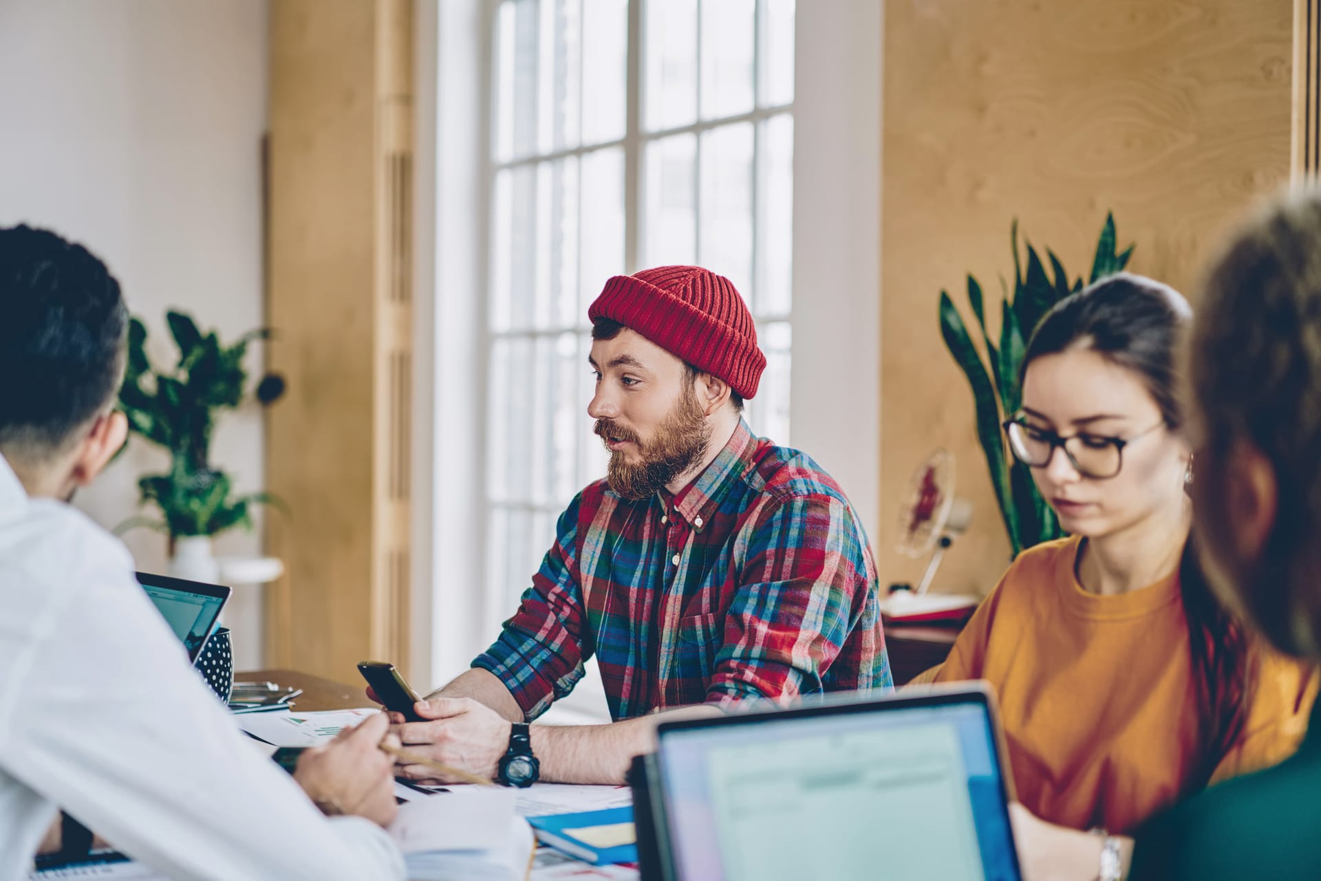 Young professionals in a bright office meeting, person in red beanie and plaid shirt speaking while others listen.