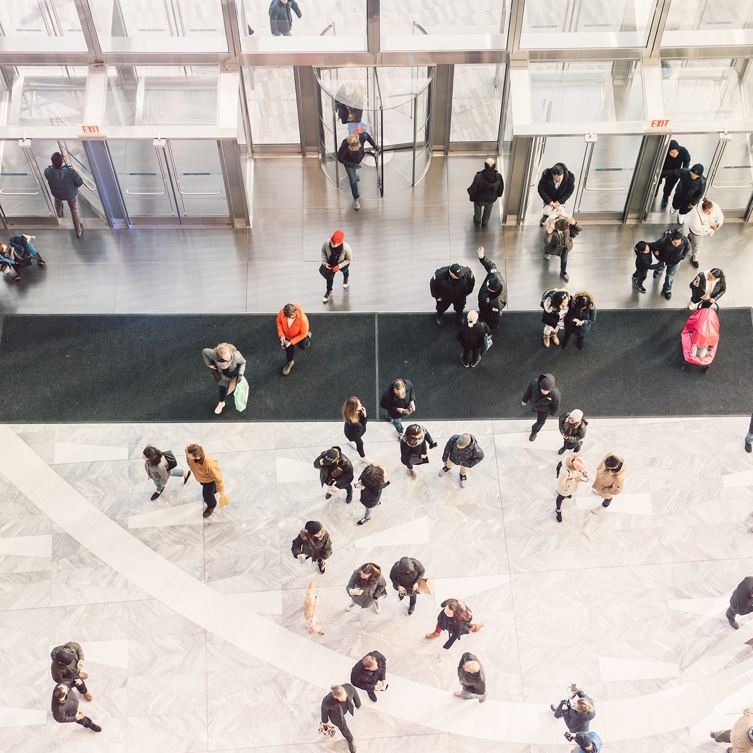 Aerial view of people walking through a modern building lobby with marble floors, glass revolving doors and exit signs.