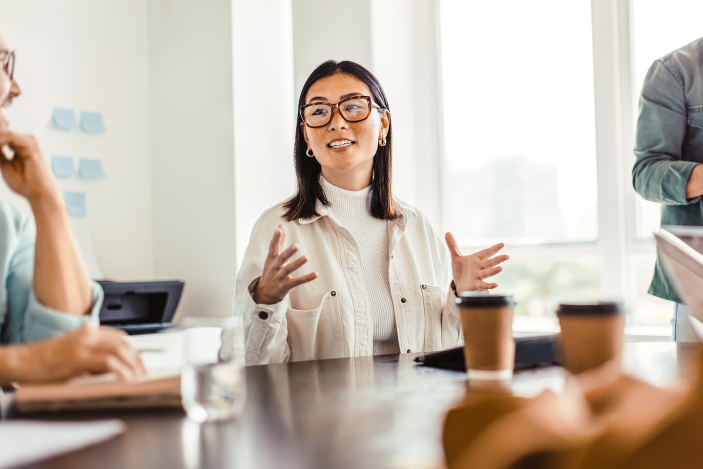Woman in glasses and white outfit speaking expressively during a meeting with coffee cups on the table in a bright office.