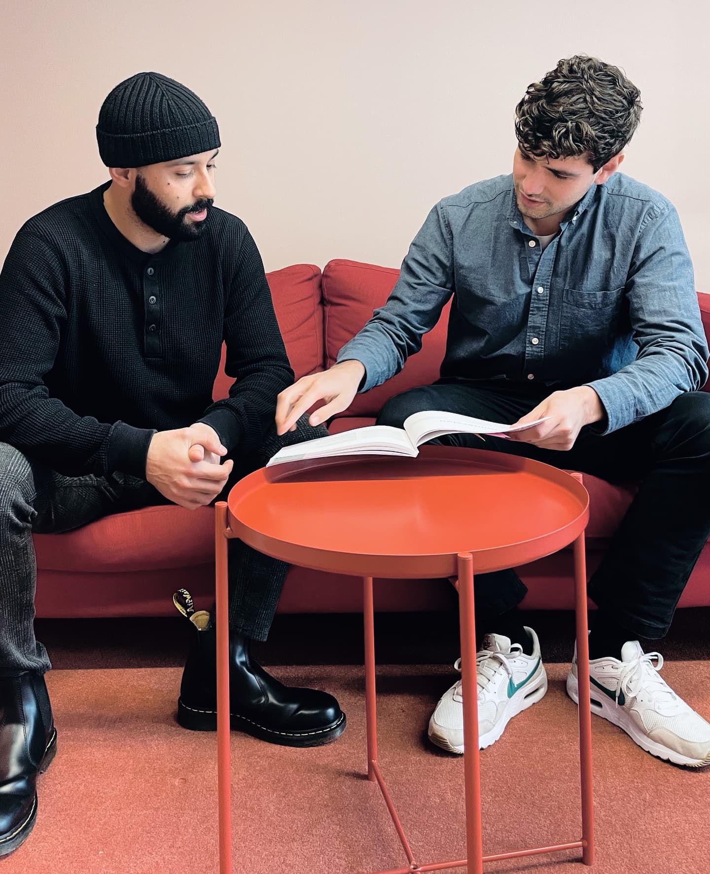 Two men sitting on a red sofa reviewing a book together, with a coral-coloured side table between them.